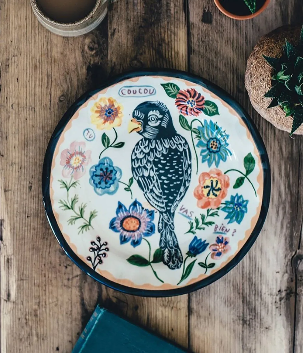 Ceramic plate with a parrot and flowers painted on it. The plate is sitting amongst potted plants, a cup of coffee and a journal.