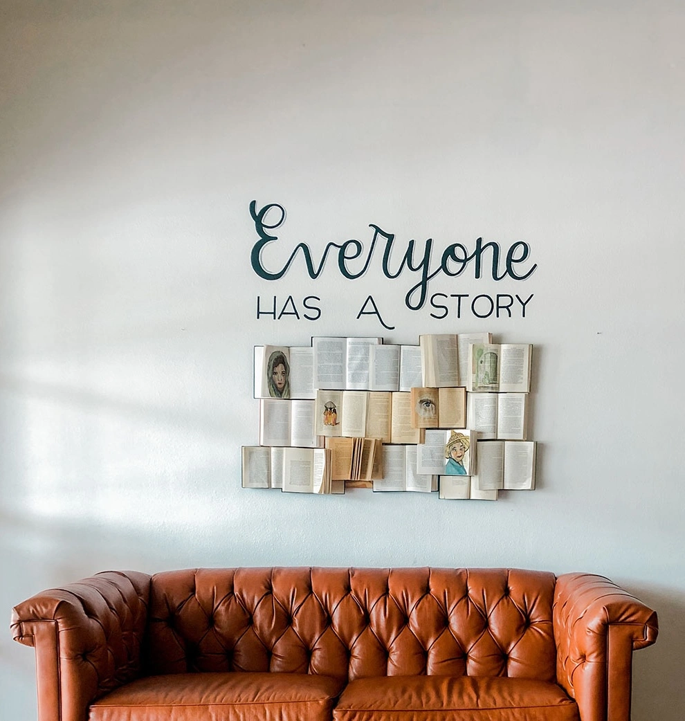 A brown leather couch against a wall with an art installation of open books and the phrase, Everyone Has a Story