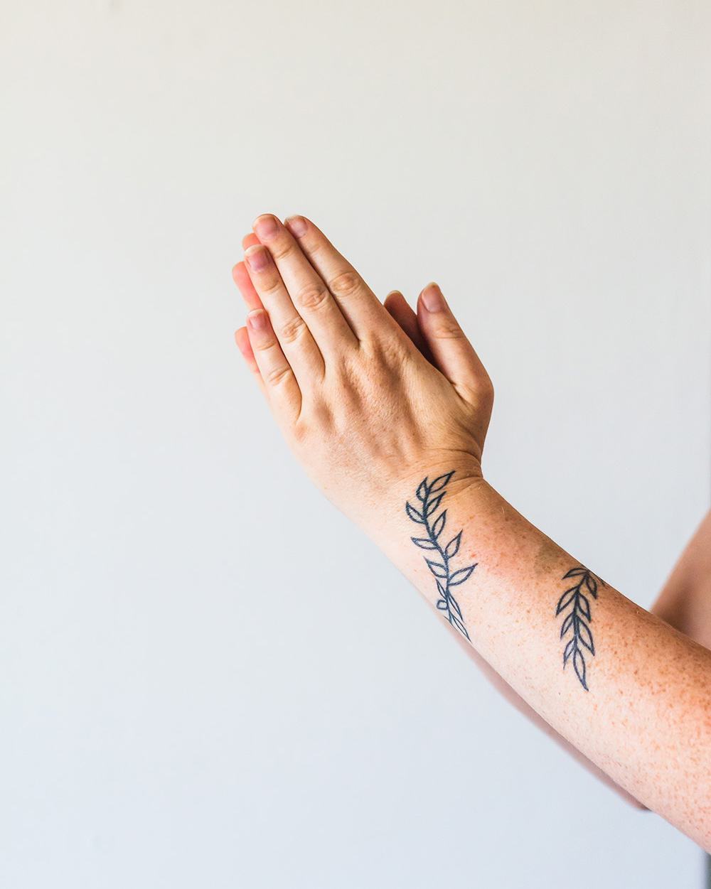 A person with their hands in anjali mudra or namaste position. They have a tattoo of leaves wrapped around their forearm.
